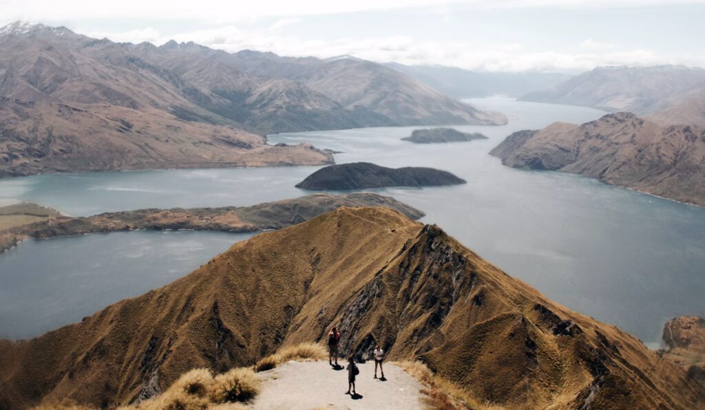 People standing on a mountain ridge above a lake and surrounding mountains.