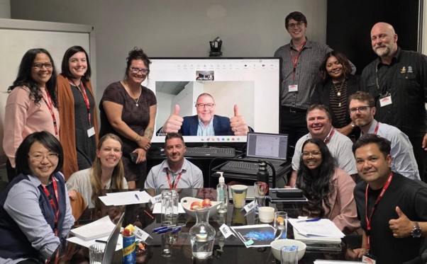 A group of professionals gather around a TV screen displaying a zoom call with a man.