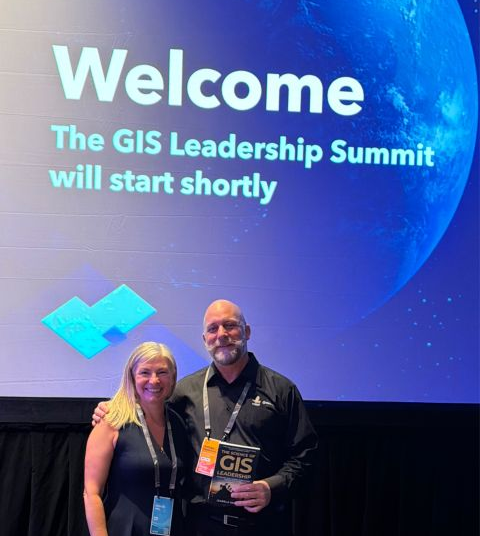 A man holding a book stands next to a woman in front of a welcome sign for The GIS Leadership Summit