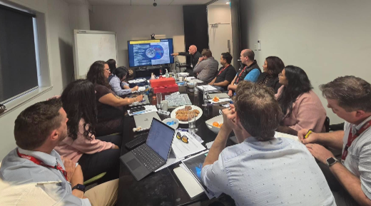 A group of professionals sit around a cluttered meeting table watching a presenter point to a TV screen.